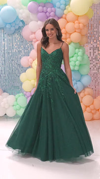 Model wearing a dark green prom gown with a fitted, embellished bodice and full tulle skirt, standing in front of pastel balloon backdrop for formal event.