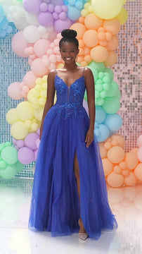 Full-length front view of the woman in a royal blue prom gown with sequined bodice, slit, and tulle skirt, smiling in front of pastel balloon backdrop.