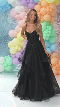 Woman in a black prom gown with beaded bodice, spaghetti straps, and voluminous tulle skirt, standing against pastel balloon backdrop